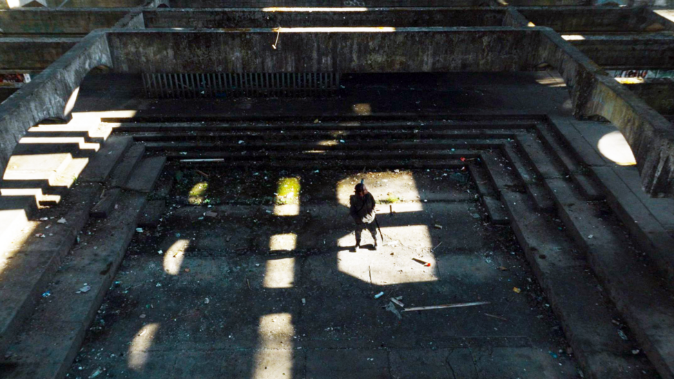 Jade McDonald walks through a concrete-clad open space that is deep in shadow.