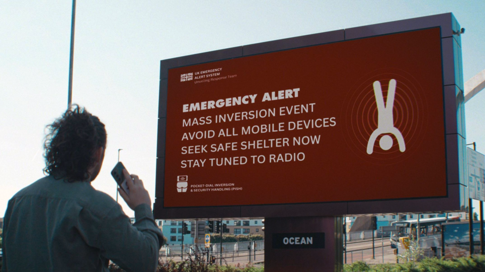 A man looks up to a large electronic sign warning of an emergency. In white letters on a red background are the words: "Emergency alert. Mass inversion event. Avoid all mobile phones. Seek safe shelter. Stay tuned to radio".