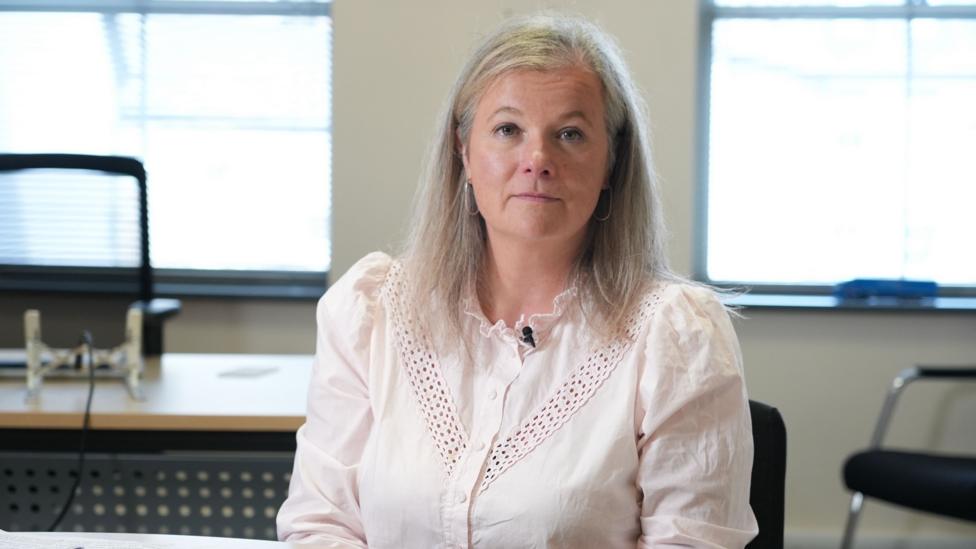 Lowri Griffiths is sitting behind a desk and is wearing a white shirt with a frilly neck. A small microphone is clipped to her collar and Lowri has shoulder length blonde/grey hair.