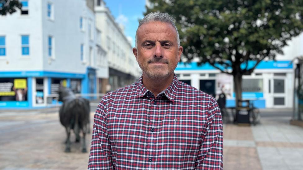 Carl is standing in a pedestrianised town square, wearing a red and blue checkered shirt. Trees, shopfronts and a sculpture are visible in the background, with a shallow depth-of-field creating a blurred backdrop.