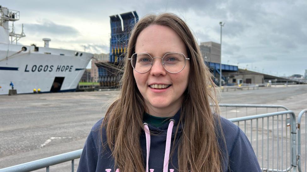 A woman in a blue and pink hoodie with long brown hair, smiling at the camera. She is wearing glasses. Behind her, the Logos Hope is visible in the background.