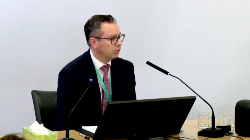 Gareth Harry from NHS England sits in front of a desk at the Civic Centre in Chelmsford where he gives evidence. He is wearing a black suit jacket and a red patterned tie. He has a green lanyard around his neck and is wearing black glasses. A box of tissues sits beside him and there is a black mic to the front of him