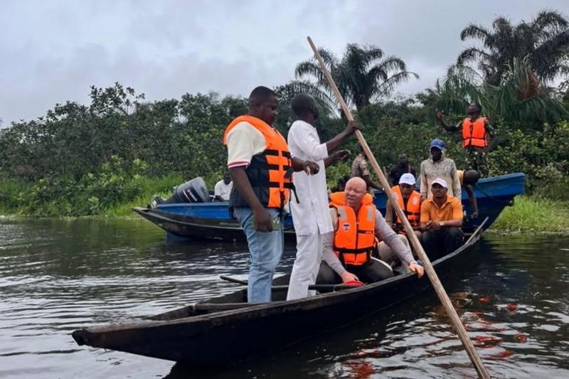 A group of people in a wooden canoe. They are wearing orange life jackets. In the background, you can see lots of greenery including palm trees