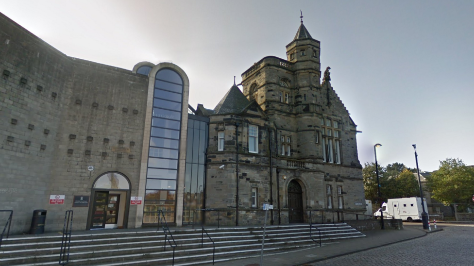 A general view of Kirkcaldy Sheriff Court. An older building made of dark stone is connected to a newer building, made of lighter stone, by several glass panels. A large brick archway is in the middle, which is also filled with glass panels.
