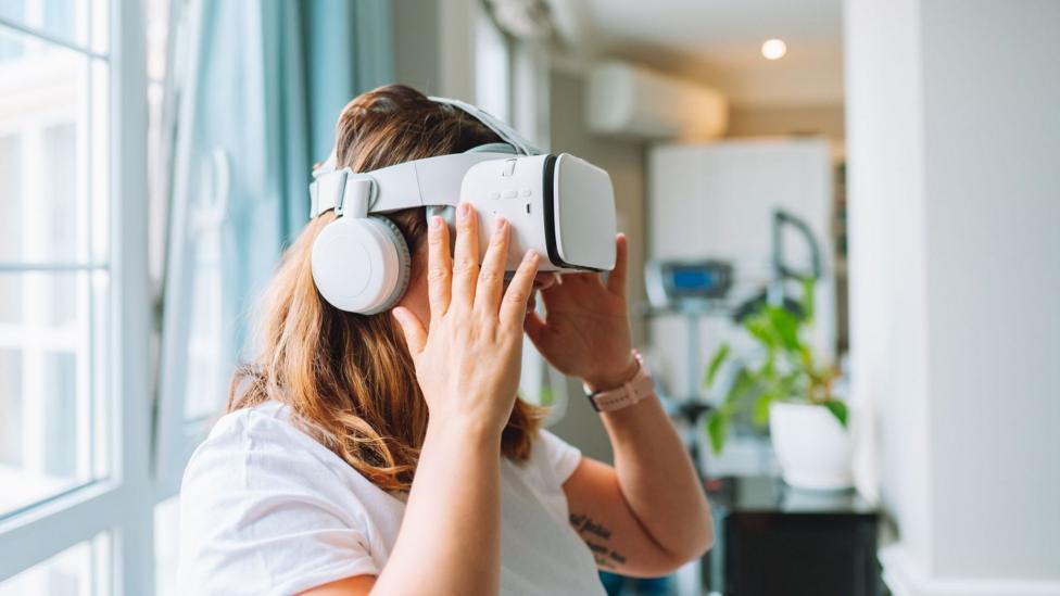 A woman with brown hair using a white virtual reality headset piece, she is wearing a white top and has her hands on the set. The background has a big window on the left and a black table with a potted plant on top on the right. 