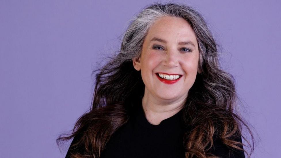 A woman with brown hair that has highlights of grey at the front smiling. She is wearing dark eyeshadow and red lipstick, and is wearing a black jumper. The background is purple. 
