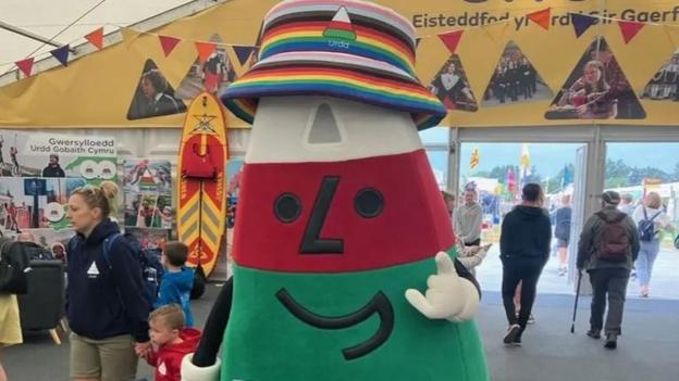 Mistar Urdd mascot. A colourful character shaped like a triangle, with a green base, red middle, and white top. He wears a rainbow bucket hat and stands in a marquee. 