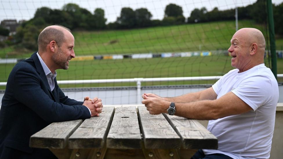 Prince William and Ross Kemp smile at each other as they sit on opposite sides of a wooden bench outside.