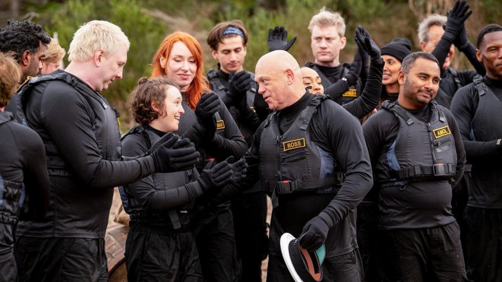 Ross Kemp and contestants from Celebrity Traitors stand together as they wear black suits and protective vests.