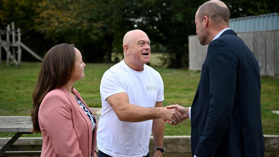 Ross Kemp and Prince William shake hands as they stand next to a woman who is wearing a pink blazer. They are standing outside.
