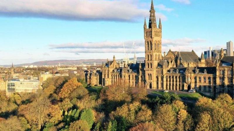Glasgow university bathed in sunlight on an autumn day. It is up on a hill with autumnal trees below and buildings in the distance