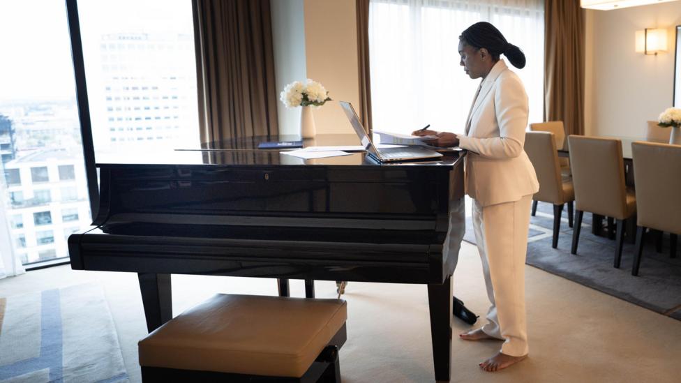 A barefoot Kemi Badenoch, dressed all in white, puts the finishing touches to her conference speech on a laptop computer resting on a grand piano, in her Birmingham hotel suite
