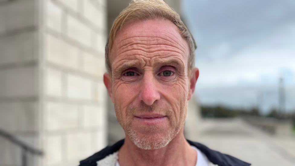 Neville Gibbons. He has short ginger hair, wearing a black hoodie.
