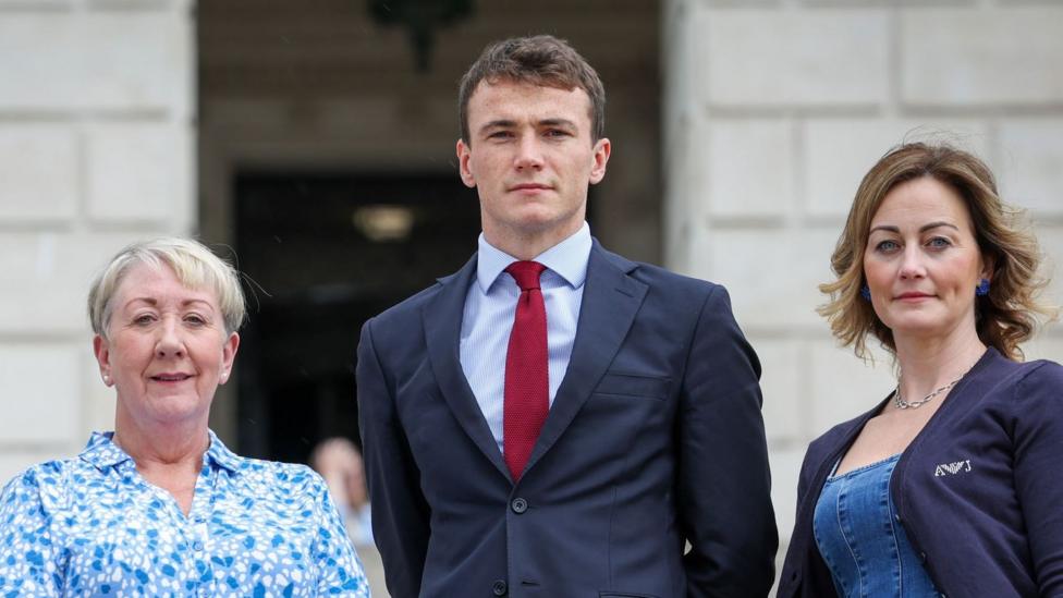 Three women and one man stand on the steps of Stormont. One woman is wearing a green blouse, another is wearing a white and blue patterned blouse, and the third woman is wearing a denim dress with a navy cardigan. The man is wearing a dark suit, blue shirt and red tie.
