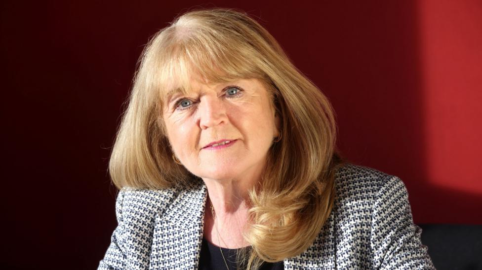 Woman with mid-length blonde hair, wearing a black top and a black and white patterned blazer.