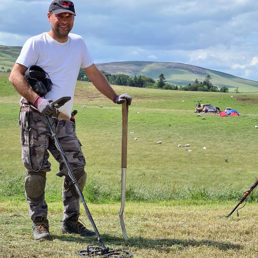 A smiling man in a cap, white t-shirt and cargo trousers is standing in a field holding a metal detector and a spade