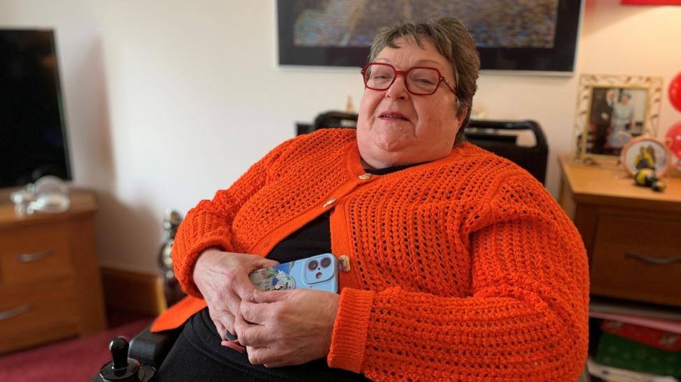 A woman in an orange cardigan holding a mobile phone. She has short brown hair and red glasses and is sitting in her living room