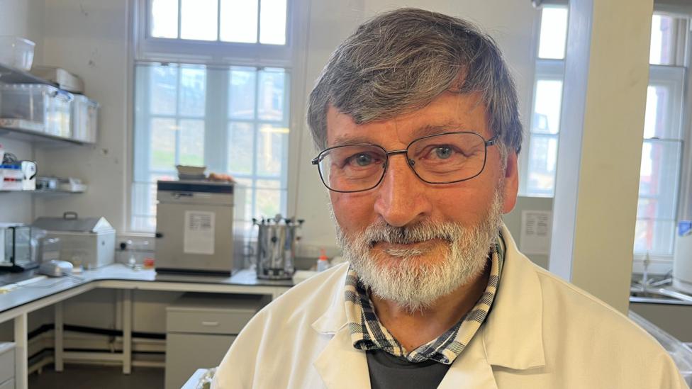 Dan Turner dressed in a white lab coatm with a navy blue checked shirt and a grey jumper. He looks at the camera in a laboratory, he has grey hair and a grey beard.