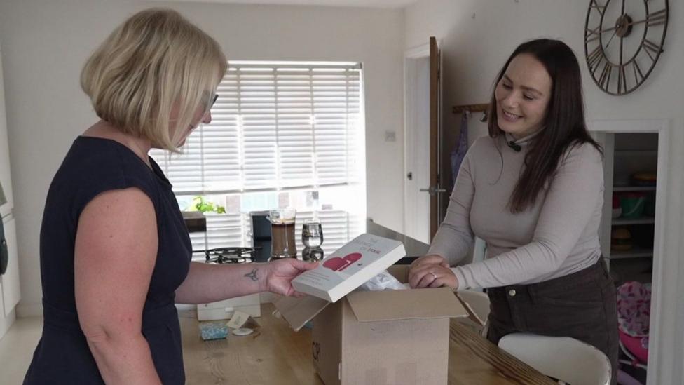 Rosanna Brown is standing in her kitchen with a brown cardboard box on the worktop. She has long brown hair over one of her shoulders. She is wearing a beige polo neck top and brown trousers. A woman with a blonde bob wearing a sleeveless dark top is holding a white book with a pink graphic on the cover.