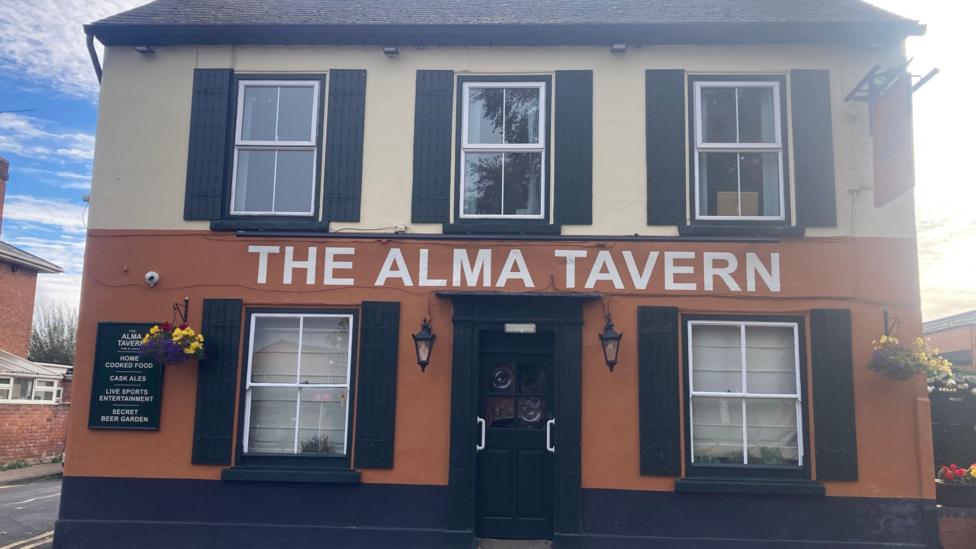 The outside of the Alma Tavern pub. The building is painted half cream, half brown, with large white writing above the black front door which spells out 'The Alma Tavern'. There are shuttered windows with four sectioned panes of glass each either side of the front door and three shuttered windows above with four sectioned panes of glass.