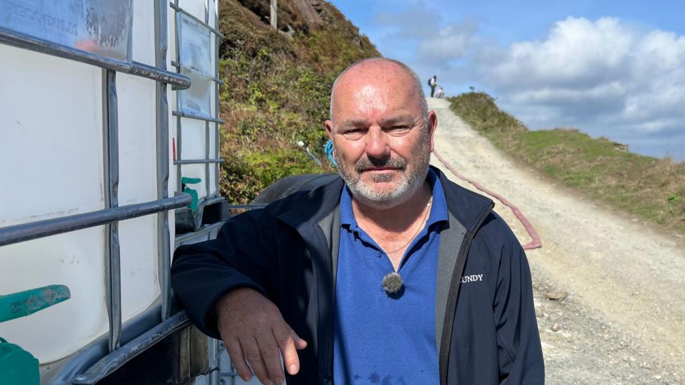 Derek Green smiling at the camera and stood next to one of the large tanks. He is wearing a blue t-shirt and a navy blue jacket. He has a short beard.