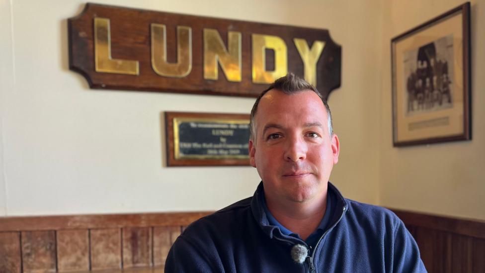 Matt Scantlebury smiling at the camera. He is wearing a blue jumper and is sat in the local pub, with a large, gold 'LUNDY' hung on the wall behind him. There is a framed image to the right and wooden wall panelling below.