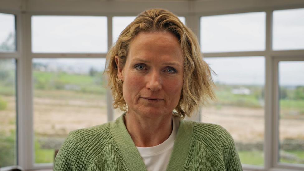 Rhianon Bragg looking at the camera. She has blue eyes and blonde hair, and has a neutral expression. She is wearing a green cardigan over a white t-shirt. She is standing in front of a window during the daytime.