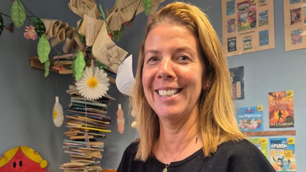 Kirsty Logan-Hall - a woman with shoulder-length blonde hair - smiles at the camera in a classroom setting. She is wearing a black top and a gold pendant necklace.