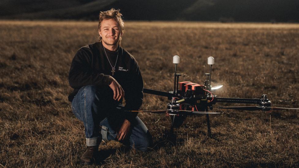 Augustus Doricko is one one knee next to a large drone. They are in a field.