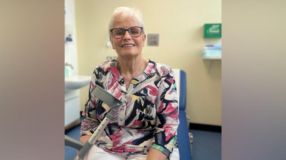 Marie Holland is sitting in a hospital room smiling and looking toward the camera. She is wearing a pink and black patterned top and white trousers, she is holding a grey crutch in one hand.
