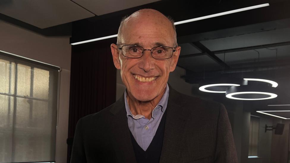 Professor Raphael Bengoa is smiling and looking into the camera. He wears dark rimmed glasses and a light blue shirt with a dark jumper and jacket. 