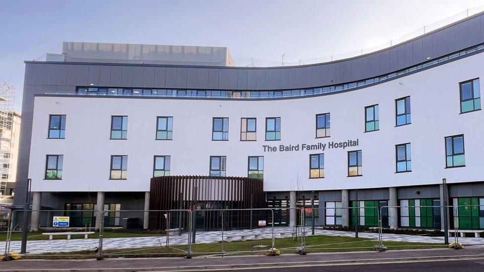 The hospital is a large, four storey white and grey building, part of the building is blue and extends from the front of the hospital.