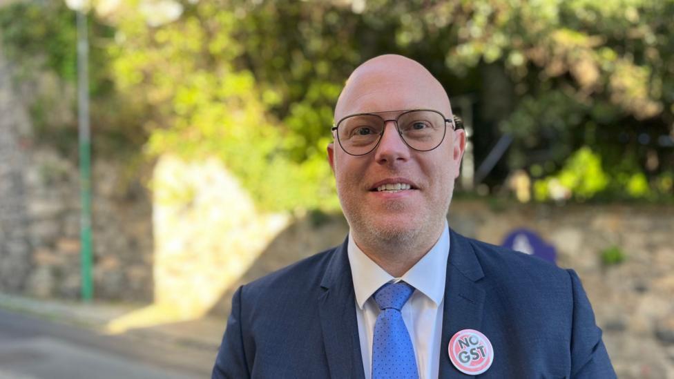 The image shows Deputy Rob Curgenven. He is a man with a bald head, wearing a blue suit and a white shirt with a blue tie. He is wearing a "No GST" badge on his left lapel.