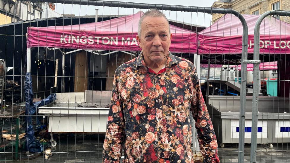 Tim Meads stands to camera with a sad expression. He is standing by his damaged stall, in front of a security gate which blocks his access. It is raining. He is wearing a flowery shirt.