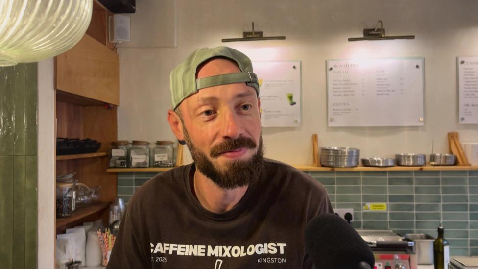 A man in a cap worn backwards looks off camera, whilst standing in a coffee shop