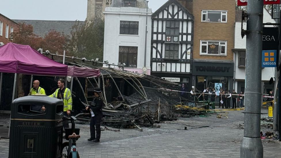 Charred and collapsed market stalls in Kingston upon Thames, with debris scattered across the marketplace as police officers and other people stand behind cordons. All Saints Church and surrounding shops can be seen in the background.