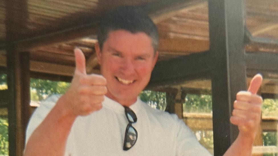 Michael Armstrong smiles and gives two thumbs up to the camera. He is clean shaven with short thick dark hair. He is wearing a white tshirt and has sunglasses hanging form the collar.