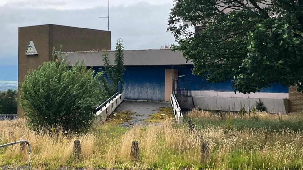 Overgrown bushes and grass are visible near a boarded up large grey and blue building.