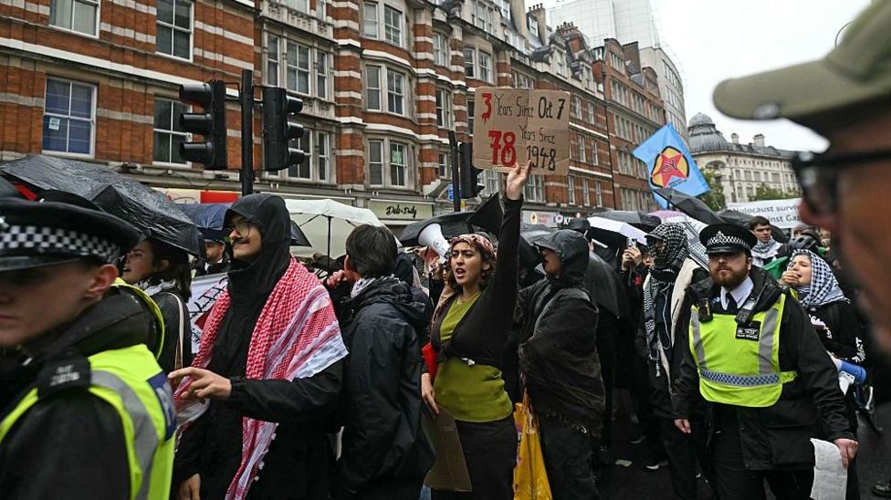 A group of people, some waving flags or holding placards, take part in a pro-Palestinian march in London. One protester can be seen holding a sign that reads "3 years since October 7, 78 years since 1948". Police can be seen escorting the march. 