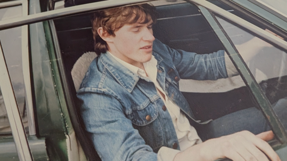 An old photo of a young man with floppy brown hair, sitting in the driver's seat of a car, with the window down and door open. He's wearing a white shirt, jeans and a blue denim jacket.