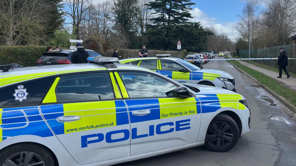 Two marked fluorescent yellow, blue and white police cars. Blue and white police tape has been put across the road to cordon it off. There are police officers at the cordon.