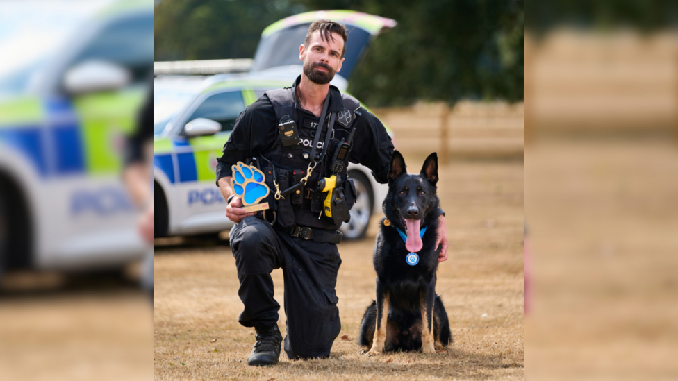 Eddie Cabrera and Frankie the German shepherd pose for a photograph. Eddie kneels using his left leg to crouch next to his partner. He holds an award in the shape of a dog's paw print, embossed in blue. He is wearing his full-black police uniform and vest. Frankie is sitting down with his ears pricked up. His large tongue is sticking out of his mouth as he sits on his hind legs. The duo are positioned on a field of dry grass, with a police car behind them.
