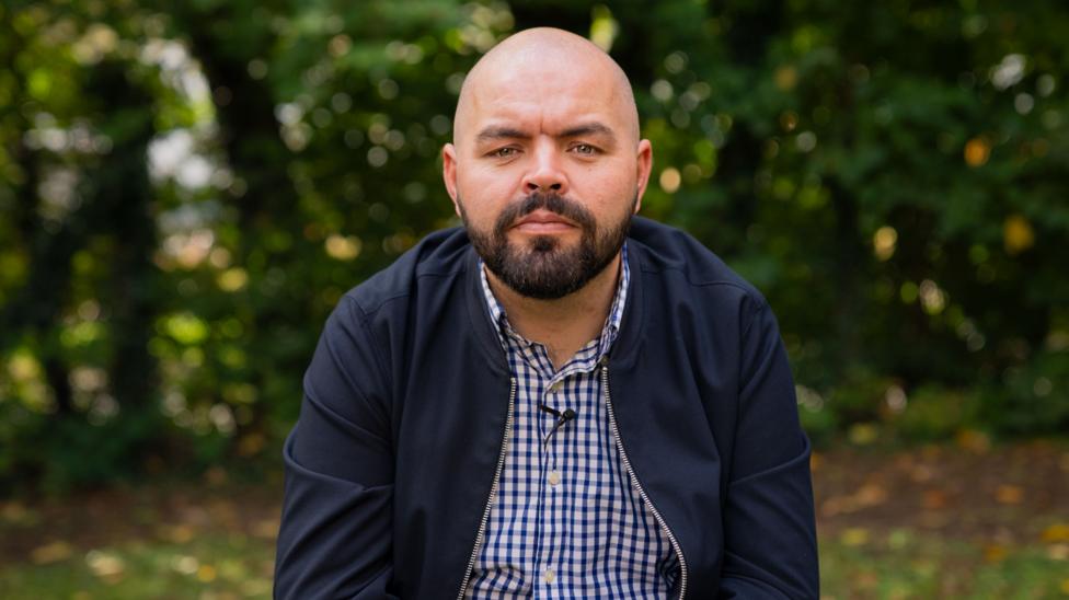 Aaron John Huxter, pictured wearing a white and navy check shirt and navy bomber jacket. He has blue eyes and a black beard. He looks directly at the camera.