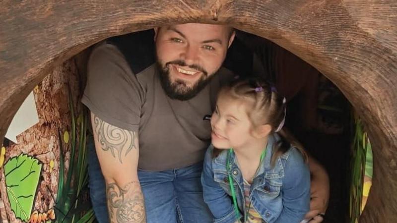 Aaron (left) smiles and is kneeling down inside a wooden tunnel. He has blue eyes and a brown beard. He leans onto his left arm, which is covered in tattoos. He wears a brown t-shirt and blue jeans. Olivia wears a denim jacket, striped t-shirt and smiles up at him.