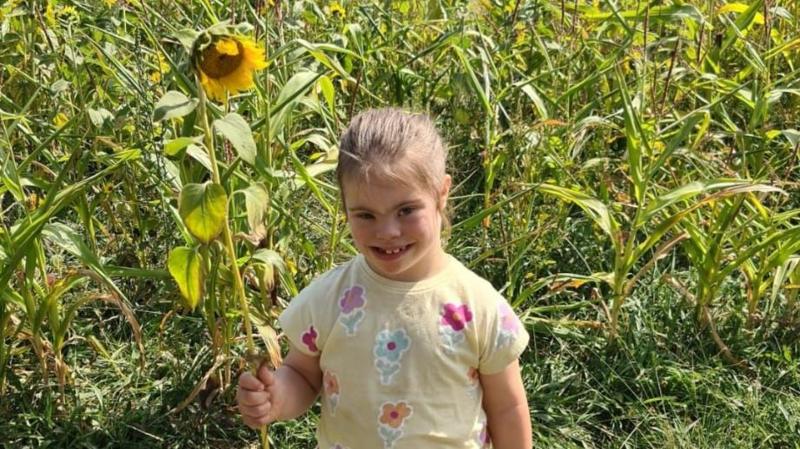 Olivia-Grace, pictured holding a large sunflower in her left hand, standing in a sunflower field. She has blue eyes and brown hair tied back in a ponytail. She wears a yellow t-shirt with flowers on it.