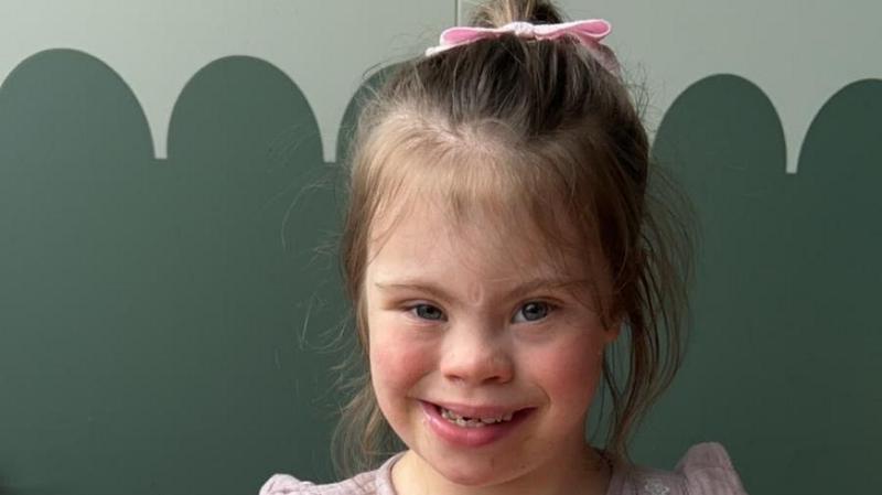Olivia-Grace pictured sat with an ice cream sundae. She wears a pink blouse with ruffled shoulders and smiles at the camera. She has blue eyes and brown hair, which is tied back into a ponytail. She has a pink bow in her ponytail. There is melted ice cream on the forefront of the table.