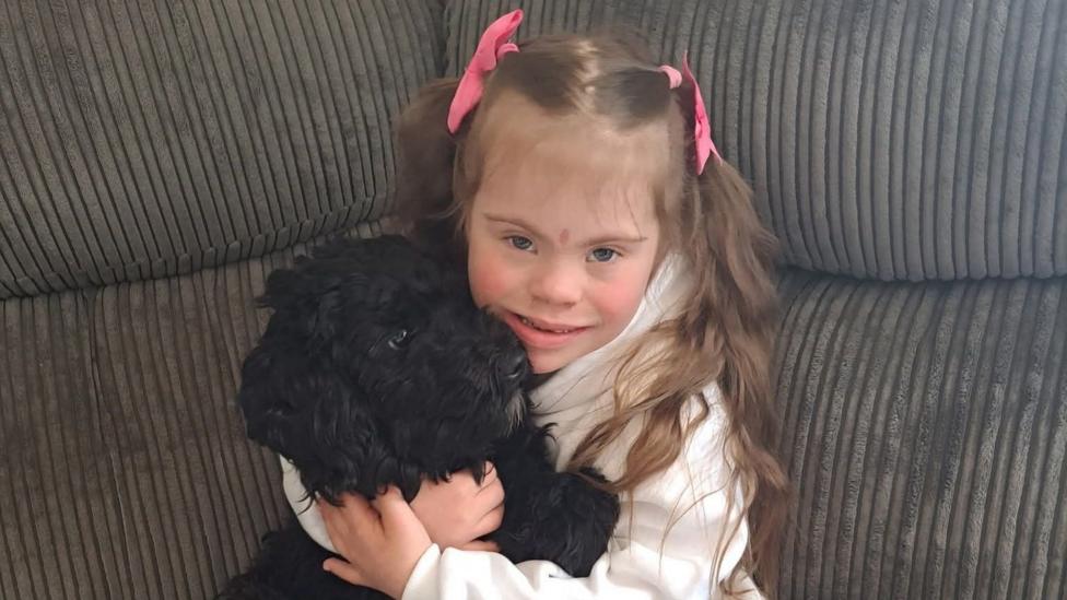 Olivia-Grace Huxter is pictured holding a black curly haired spaniel. She cradles the dog in her arms and smiles directly at the camera. She has blue eyes and brown hair which is styled in two bunches, tied with pink ribbons.