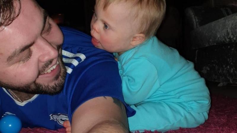 Aaron (left) lies on the floor and holds his phone in his left arm, which is in the centre of the photo. Olivia-Grace is just months old and leans on Aaron's arm, looking at him with a smile. She wears a bright blue babygrow. Aaron has blue eyes and dark brown hair.