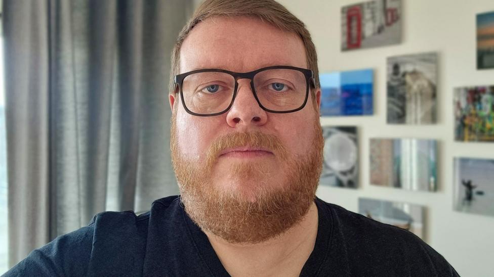 A man with a ginger beard wearing dark glasses and a dark blue t-shirt is at the camera