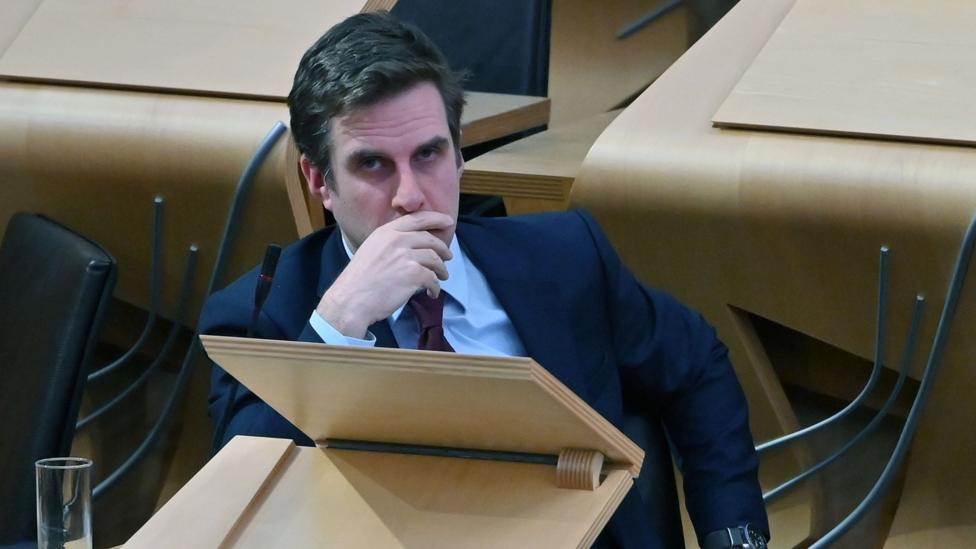 Daniel Johnson sitting behind a desk in the chamber of the Scottish Parliament. He has short black hair and is wearing a dark blue suit, light blue shirt and a tie. His right hand is covering his mouth.
